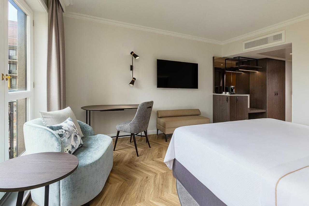 מלון The Westin Grand ברלין מראה חיצוני תמונה The photo shows a modern hotel room. It features a large bed with white linens, a stylish chair and a small round table to one side. There is a flat-screen television mounted on the wall across from the bed. In the background, there's a kitchenette a