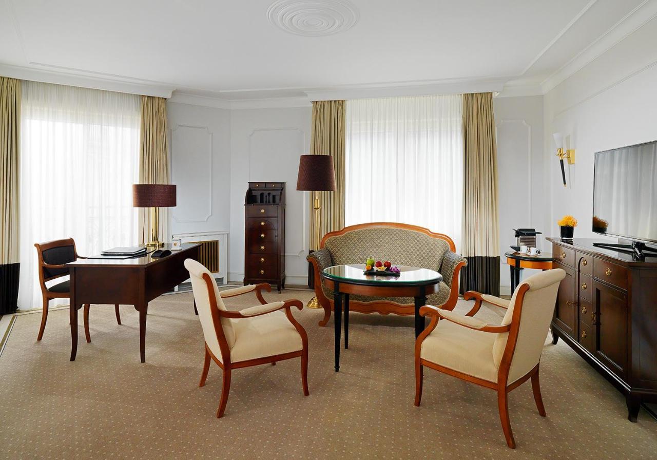 מלון The Westin Grand ברלין מראה חיצוני תמונה The photo shows a stylish hotel room interior. It features a cozy sitting area with a curved sofa and two light-colored upholstered chairs, all arranged around a small circular table. A dark wooden desk with a lamp and a vintage-style wooden cabinet 