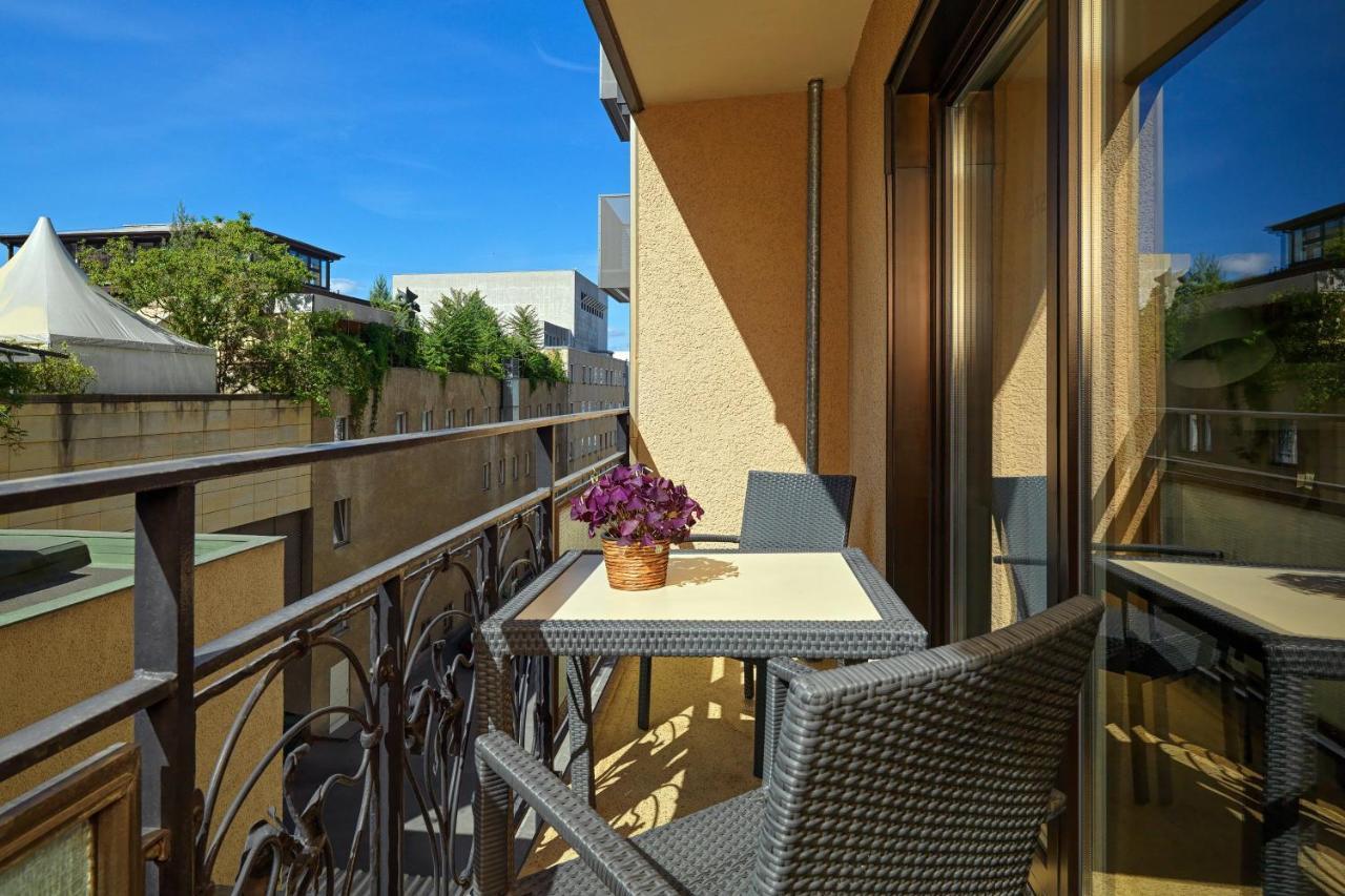 מלון The Westin Grand ברלין מראה חיצוני תמונה The photo depicts a balcony featuring a small table and two chairs. The table is set with a potted purple flower, adding a touch of decoration. The balcony has a wrought iron railing and glass walls, allowing for outdoor views. The background shows g
