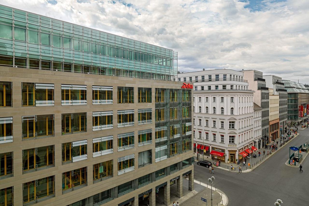 מלון The Westin Grand ברלין מראה חיצוני תמונה The photo shows an urban scene featuring a modern building on the left side, which has a glass facade and a large entrance, suggesting it may be a commercial or office structure. Adjacent to it, there is another building with traditional architecture