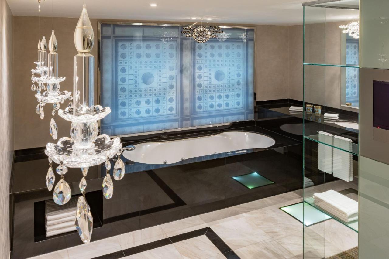 מלון The Westin Grand ברלין מראה חיצוני תמונה The photo shows a modern and luxurious bathroom. It features a large, oval bathtub that is elegantly positioned in front of large windows with patterned coverings. The room has a chic color scheme of black, white, and light gray, with marble flooring