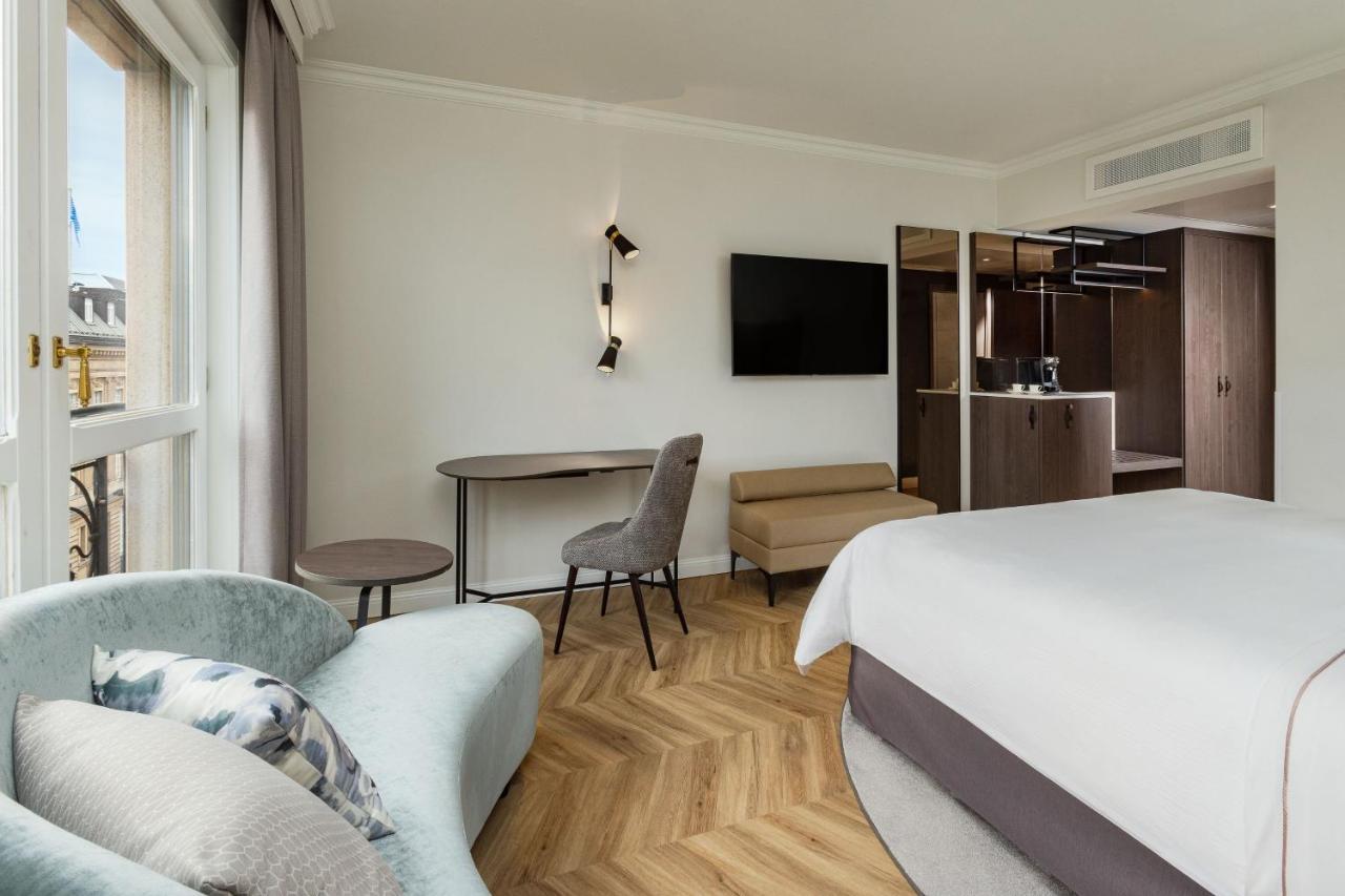 מלון The Westin Grand ברלין מראה חיצוני תמונה The photo shows a modern hotel room featuring a stylish and contemporary design. There is a comfortable bed with a light-colored bedding set, and a small sofa with a decorative pillow nearby. A round table and a gray chair are positioned in the room,