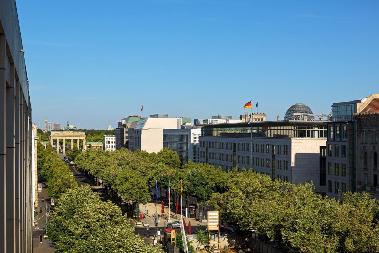 מלון The Westin Grand ברלין מראה חיצוני תמונה The photo shows a view of a cityscape with a clear blue sky. In the foreground, there is a broad avenue lined with trees. On one side of the avenue, you can see modern buildings, some of which have flags flying. Notably, there's a flag of Germany pro