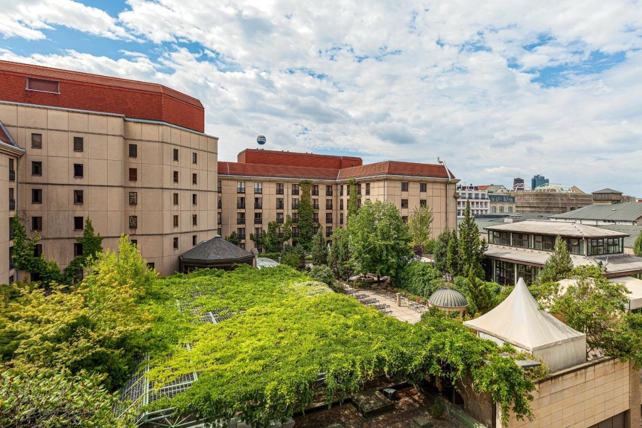 מלון The Westin Grand ברלין מראה חיצוני תמונה The photo shows a view of a building complex, likely a hotel or similar establishment, with a prominent red roof. In the foreground, there is a lush green area with a lot of vegetation, including trees and vines. There appears to be a tent structure 