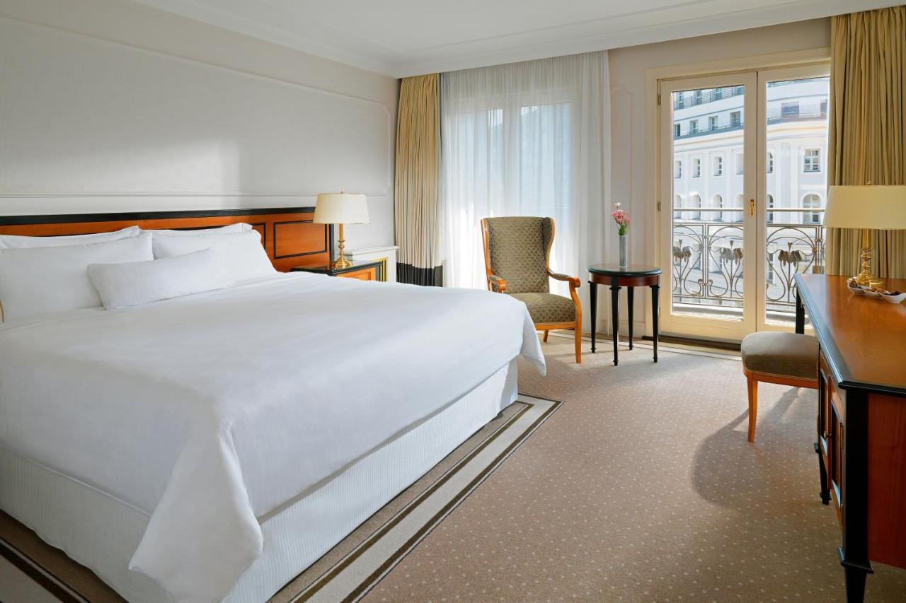מלון The Westin Grand ברלין מראה חיצוני תמונה The photo shows a hotel room featuring a large bed with white linens, a bedside lamp, and a small armchair. There is a table next to the chair, and large windows with sheer curtains allow natural light to fill the space. The decor is simple and elega
