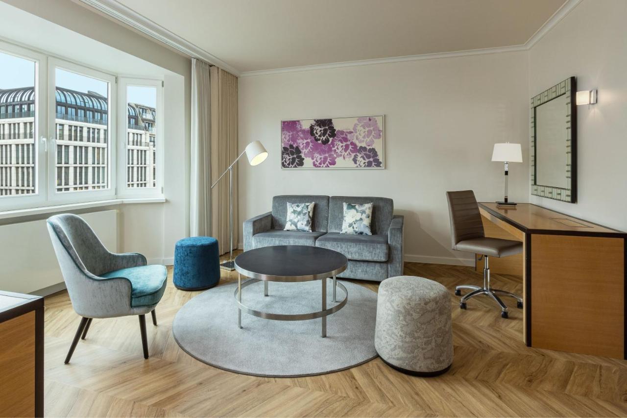 מלון The Westin Grand ברלין מראה חיצוני תמונה The photo shows a modern and stylish living area. It features a gray sofa with decorative pillows, a round coffee table in the center, and a circular rug underneath. There are two different types of seating: a blue accent chair and a small gray stool
