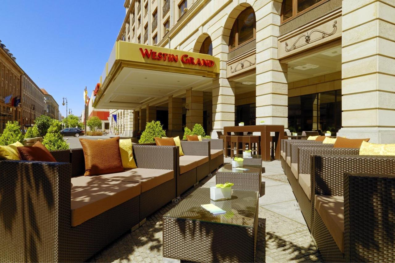 מלון The Westin Grand ברלין מראה חיצוני תמונה The photo shows an exterior view of a hotel with a prominent sign that reads "Westin Grand." In the foreground, there is a stylish outdoor seating area featuring brown wicker furniture, including couches and coffee tables. The seating is accented wit