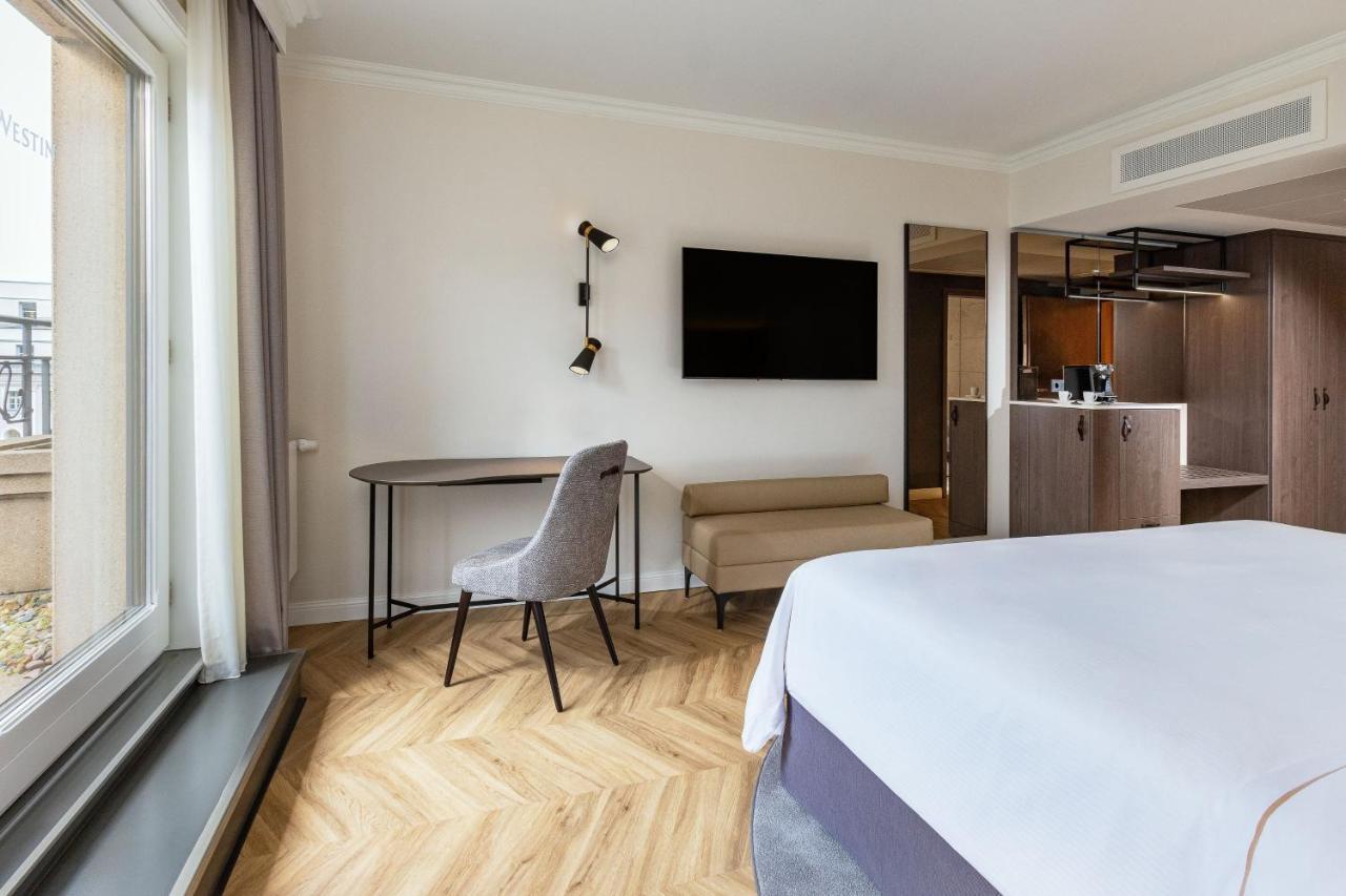 מלון The Westin Grand ברלין מראה חיצוני תמונה The photo shows a modern hotel room featuring a light-colored interior. There is a bed with crisp white linens, a small seating area with a light-colored sofa, and a round table with a chair. A large flat-screen TV is mounted on the wall, and there i