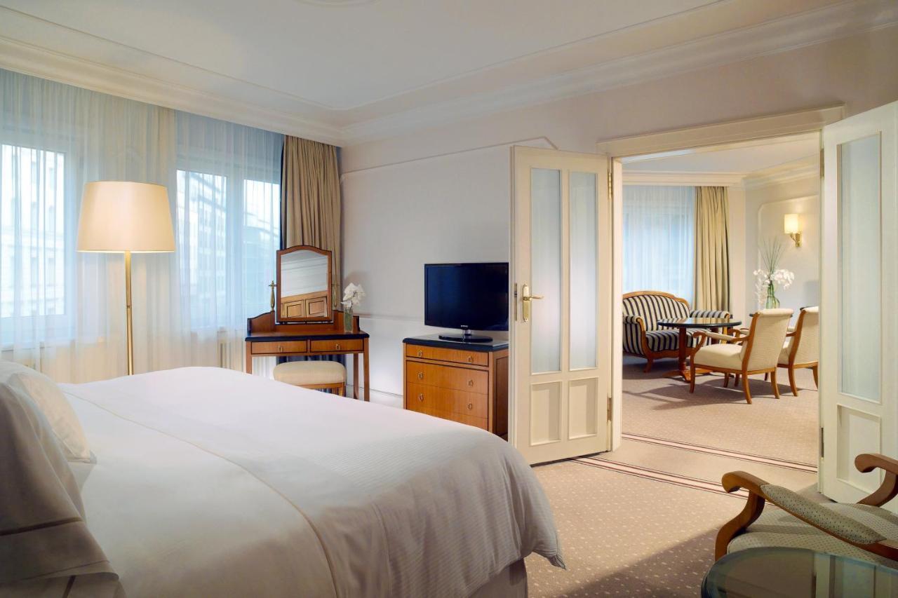 מלון The Westin Grand ברלין מראה חיצוני תמונה The photo shows a luxurious hotel room. In the foreground, there is a neatly made bed with white linens. To the left, there's a stylish floor lamp illuminating the space. Opposite the bed, there's a television on a wooden cabinet, and a vanity table 