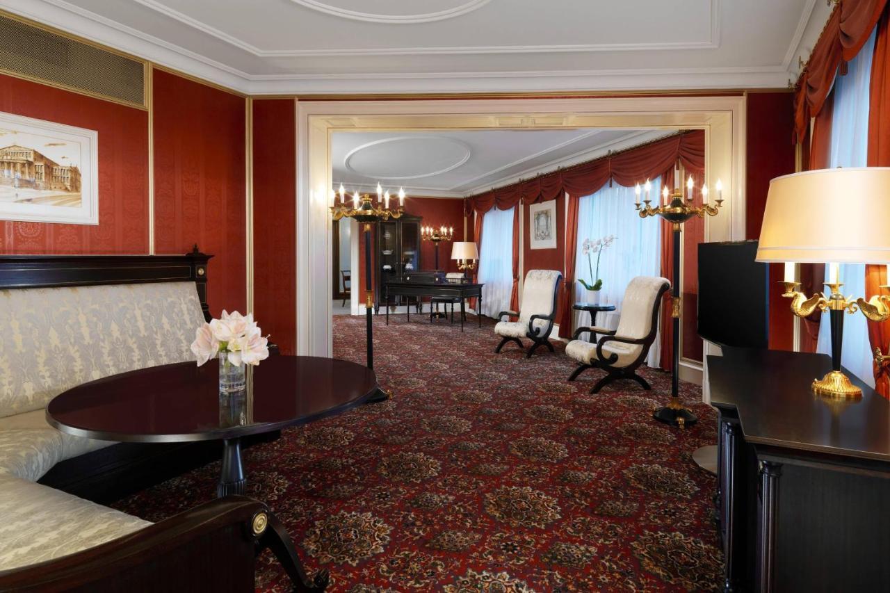 מלון The Westin Grand ברלין מראה חיצוני תמונה The photo shows an elegantly decorated hotel suite or living space. The interior features rich red wallpaper and a plush patterned carpet, creating a warm and inviting atmosphere. There's a round table with a vase of flowers in the foreground, and tw