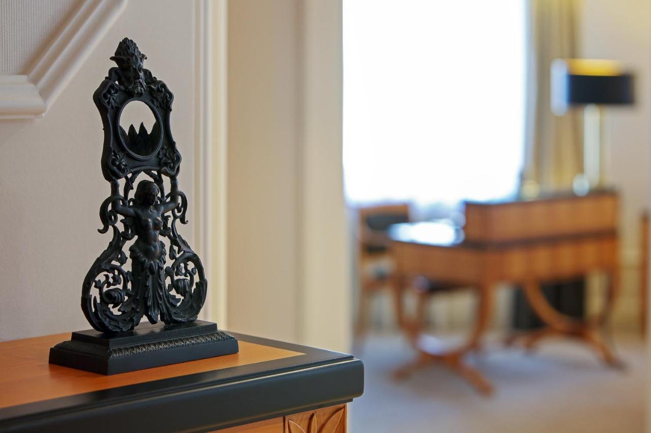 מלון The Westin Grand ברלין מראה חיצוני תמונה The photo shows a decorative black object, possibly a sculpture or a stand, placed on a surface. The object features intricate designs, including a central figure and ornate detailing. In the background, there is a blurred view of a room with light-c