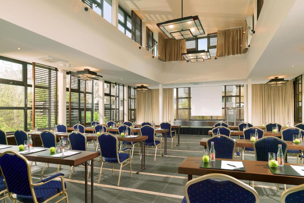 מלון The Westin Grand ברלין מראה חיצוני תמונה The photo shows a spacious, well-lit conference room designed for meetings or events. It features several rows of wooden tables arranged with blue upholstered chairs. The room has large windows adorned with curtains, allowing natural light to enter. 