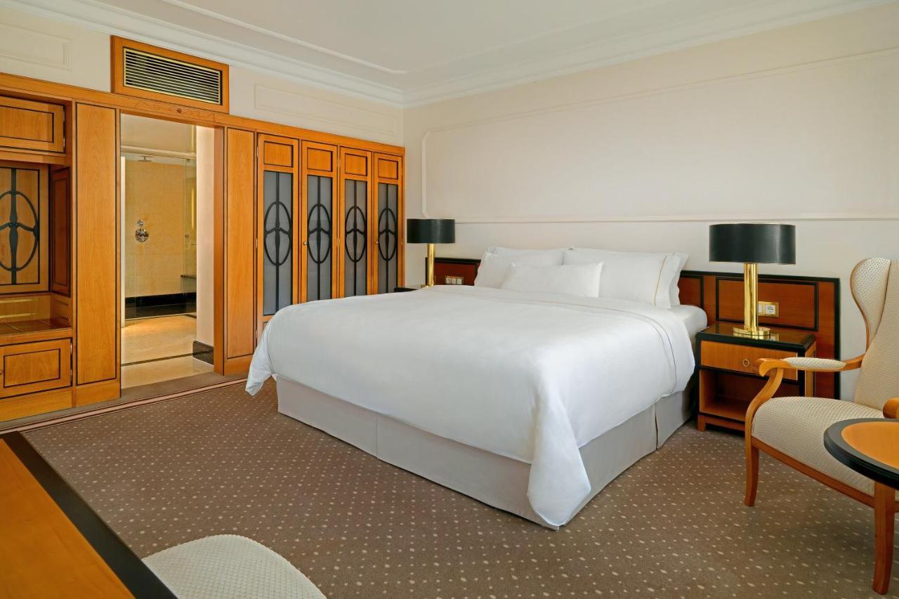 מלון The Westin Grand ברלין מראה חיצוני תמונה The photo shows a well-furnished hotel room. There is a large, neatly made bed with white bedding in the center. Two stylish bedside lamps are located on either side of the bed. To the left, there is a wooden wardrobe with glass doors. There is also 