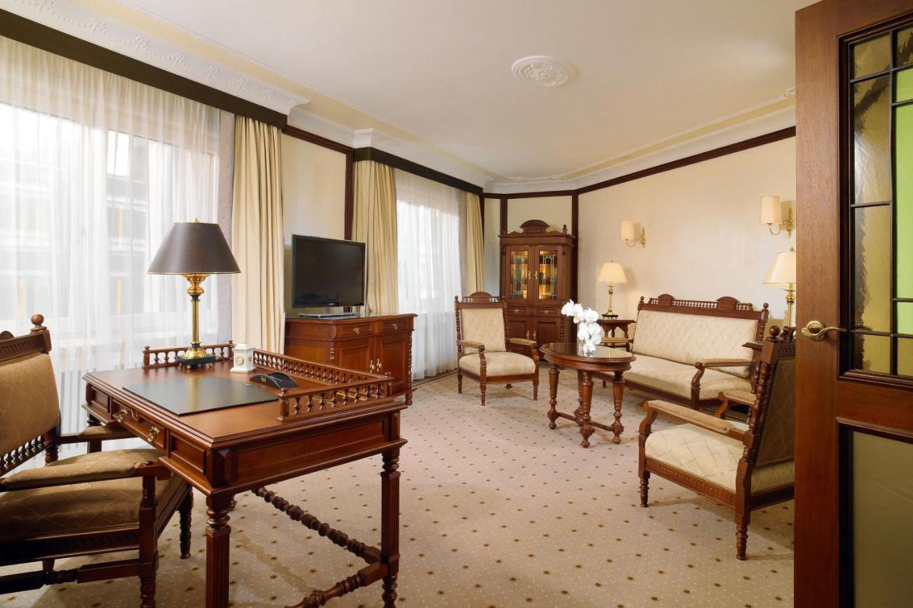 מלון The Westin Grand ברלין מראה חיצוני תמונה The photo shows an elegant room with a classic design. It features a seating area with upholstered chairs and a sofa, all arranged around a coffee table. A wooden desk with a lamp is positioned nearby, and there is a television on a cabinet. Soft cur