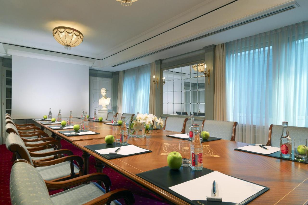 מלון The Westin Grand ברלין מראה חיצוני תמונה The photo depicts a stylish and well-lit conference room. In the center, there is a large wooden table arranged with several items, including notepads and pens at each seat, along with green apples and bottles of water. The room features elegant furn