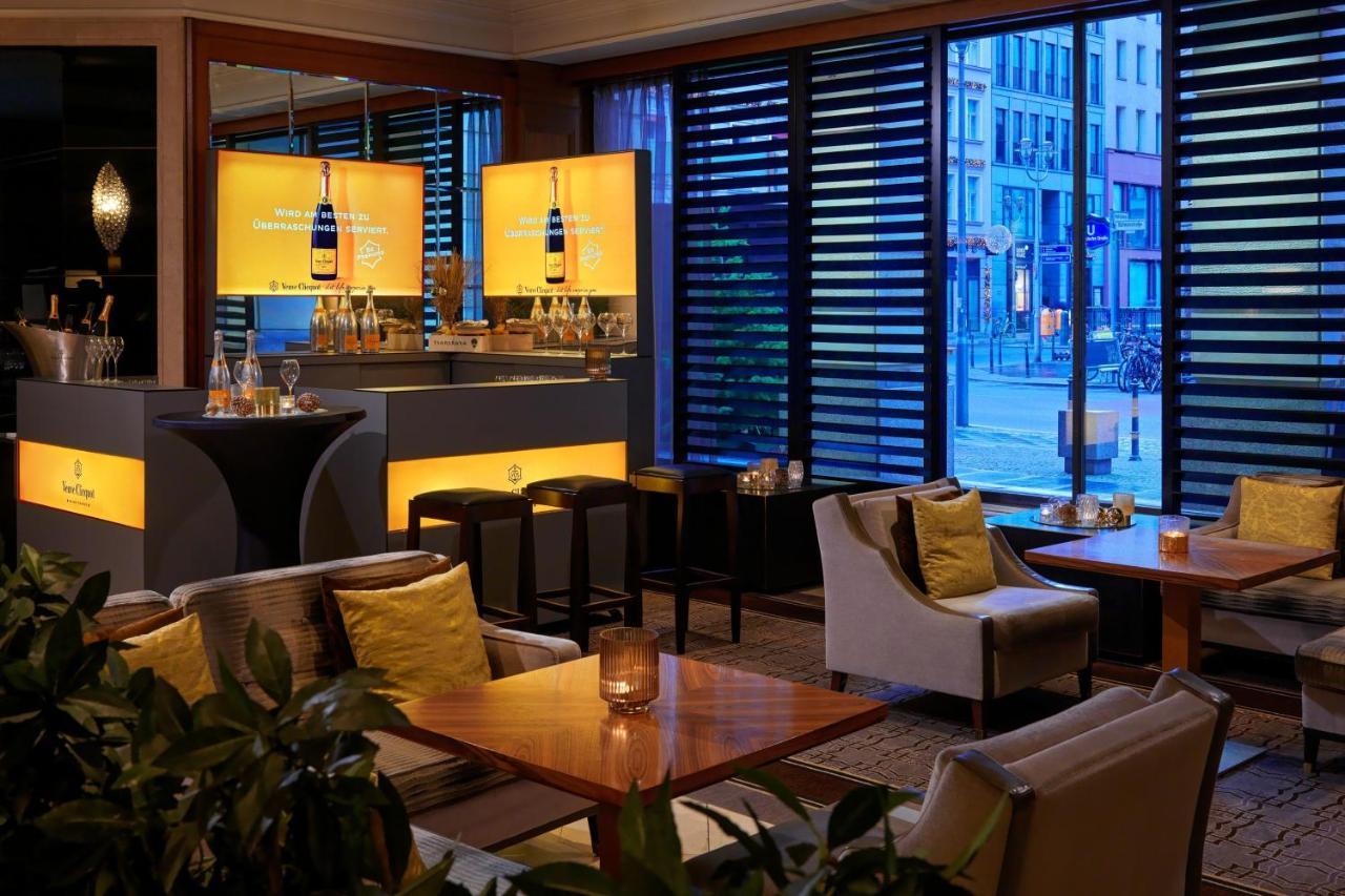מלון The Westin Grand ברלין מראה חיצוני תמונה The photo shows a modern bar or lounge area with a contemporary design. There are several tables and comfortable seating arranged around the space, featuring cozy chairs and light-colored cushions. The bar area is well-lit, with stylish displays illu