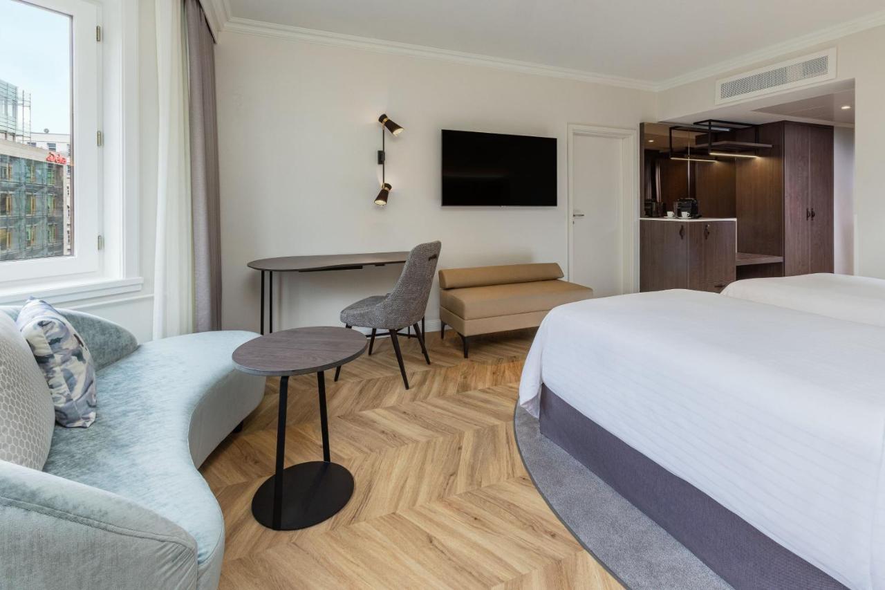 מלון The Westin Grand ברלין מראה חיצוני תמונה The photo shows a modern hotel room. It features a double bed with a light-colored comforter and decorative pillows. There's a small sofa in a soft blue color, accompanied by a round, dark table. A stylish chair is positioned near the table, and a wa