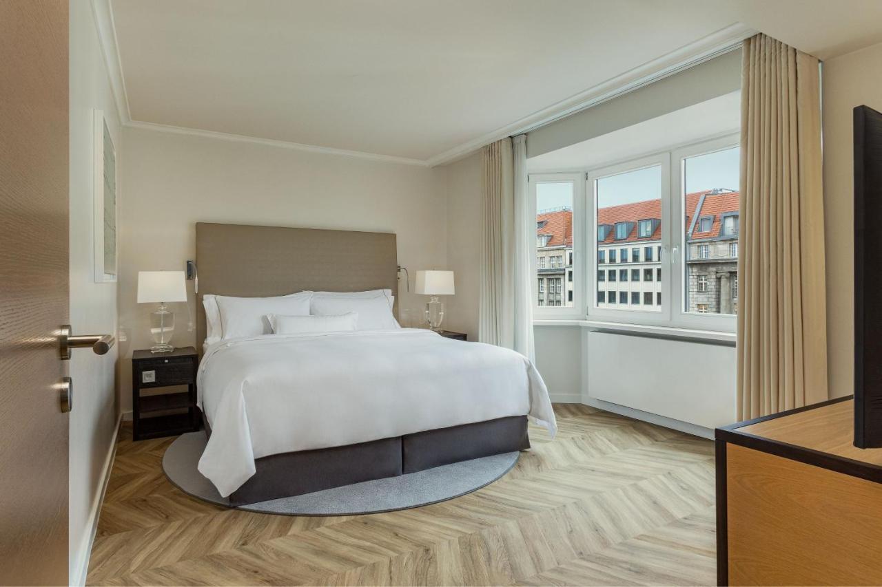 מלון The Westin Grand ברלין מראה חיצוני תמונה The photo shows a modern, elegantly furnished hotel room. It features a spacious bed with a gray upholstered headboard and crisp white linens. There are two bedside tables with lamps on either side of the bed. The flooring is wood with a herringbone 