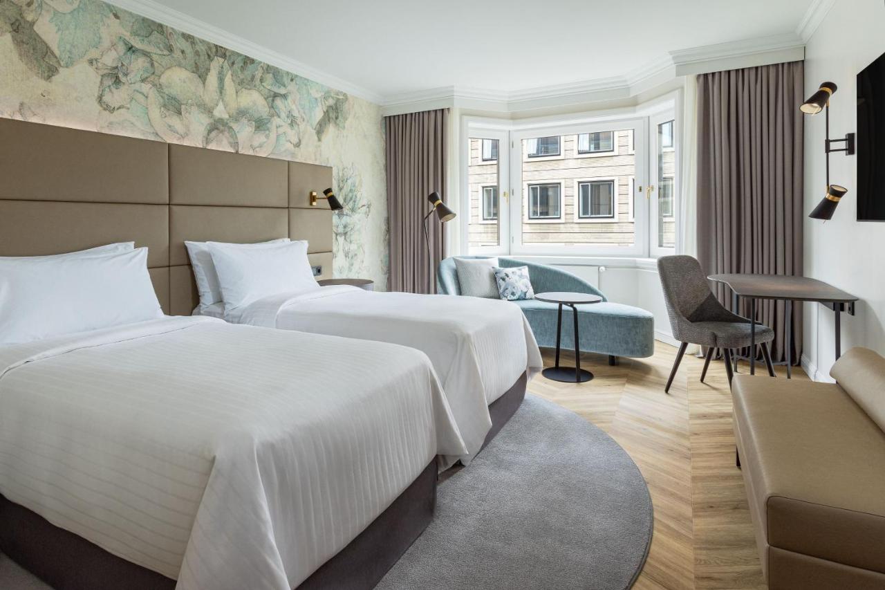 מלון The Westin Grand ברלין מראה חיצוני תמונה The photo shows a modern hotel room featuring two twin beds with white linens arranged neatly. The walls are adorned with a subtle floral wallpaper, creating a serene atmosphere. There's a small seating area near a large window, which allows for natu