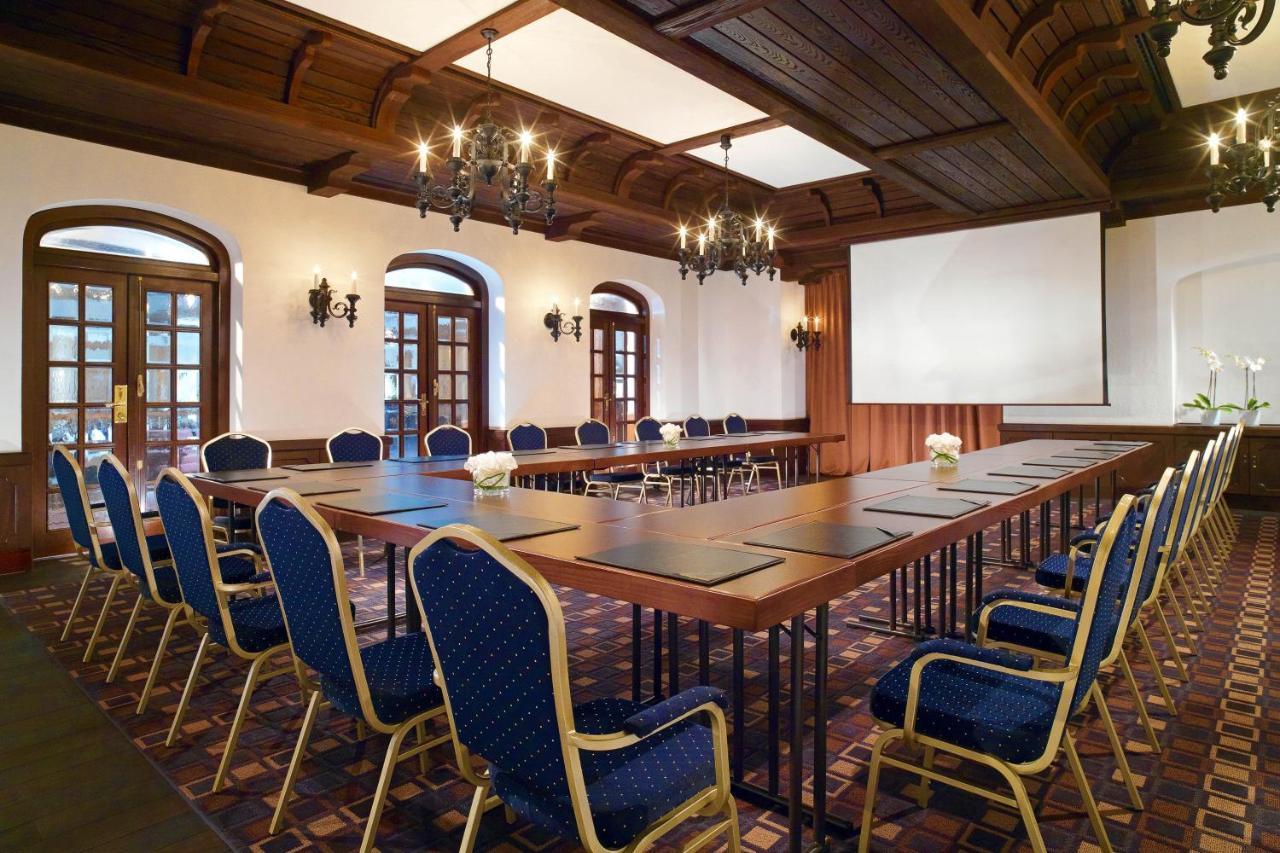 מלון The Westin Grand ברלין מראה חיצוני תמונה The photo shows a conference room with an elegant design. There is a long rectangular table set up in the center, surrounded by upholstered chairs with blue fabric. The room features wooden ceiling beams and decorative lighting fixtures, including ch