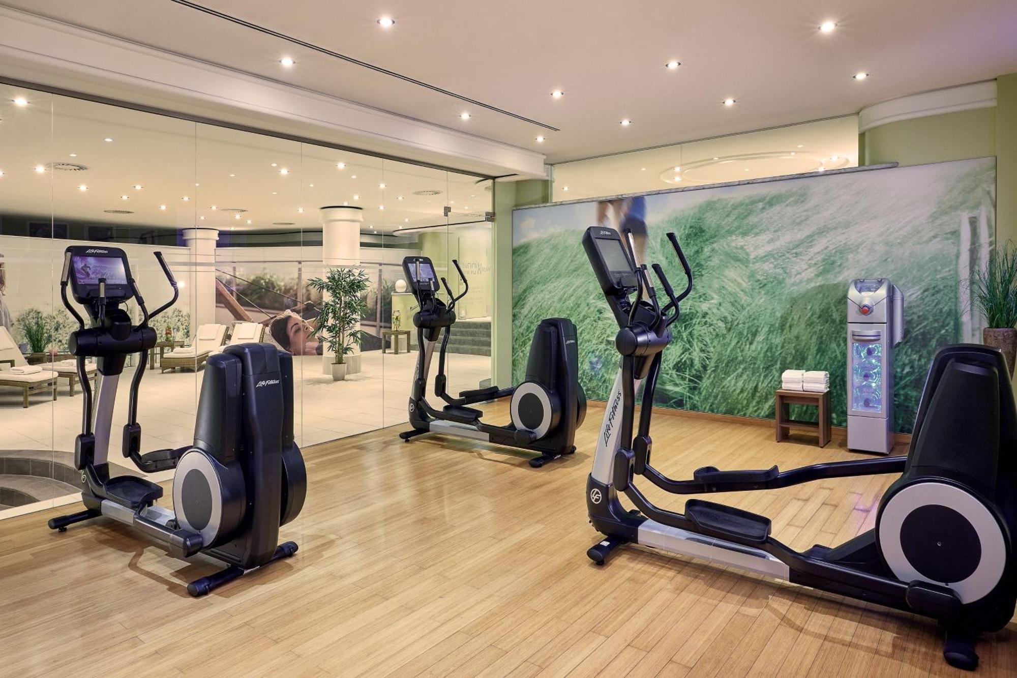 מלון The Westin Grand ברלין מראה חיצוני תמונה The photo shows a fitness area equipped with several elliptical machines positioned on a hardwood floor. There is a glass wall in the background that allows a view into another room, which appears to be a lounge or common area with plants and seating
