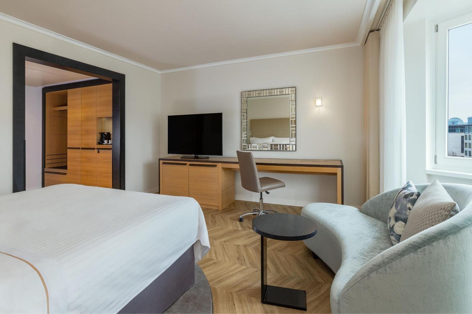 מלון The Westin Grand ברלין מראה חיצוני תמונה The photo shows a modern hotel room that features several elements:

1. **Bed**: A neatly made bed with a light-colored duvet and white sheets.
2. **Furniture**: There is a small round table next to a contemporary gray sofa, and a desk with a chair p