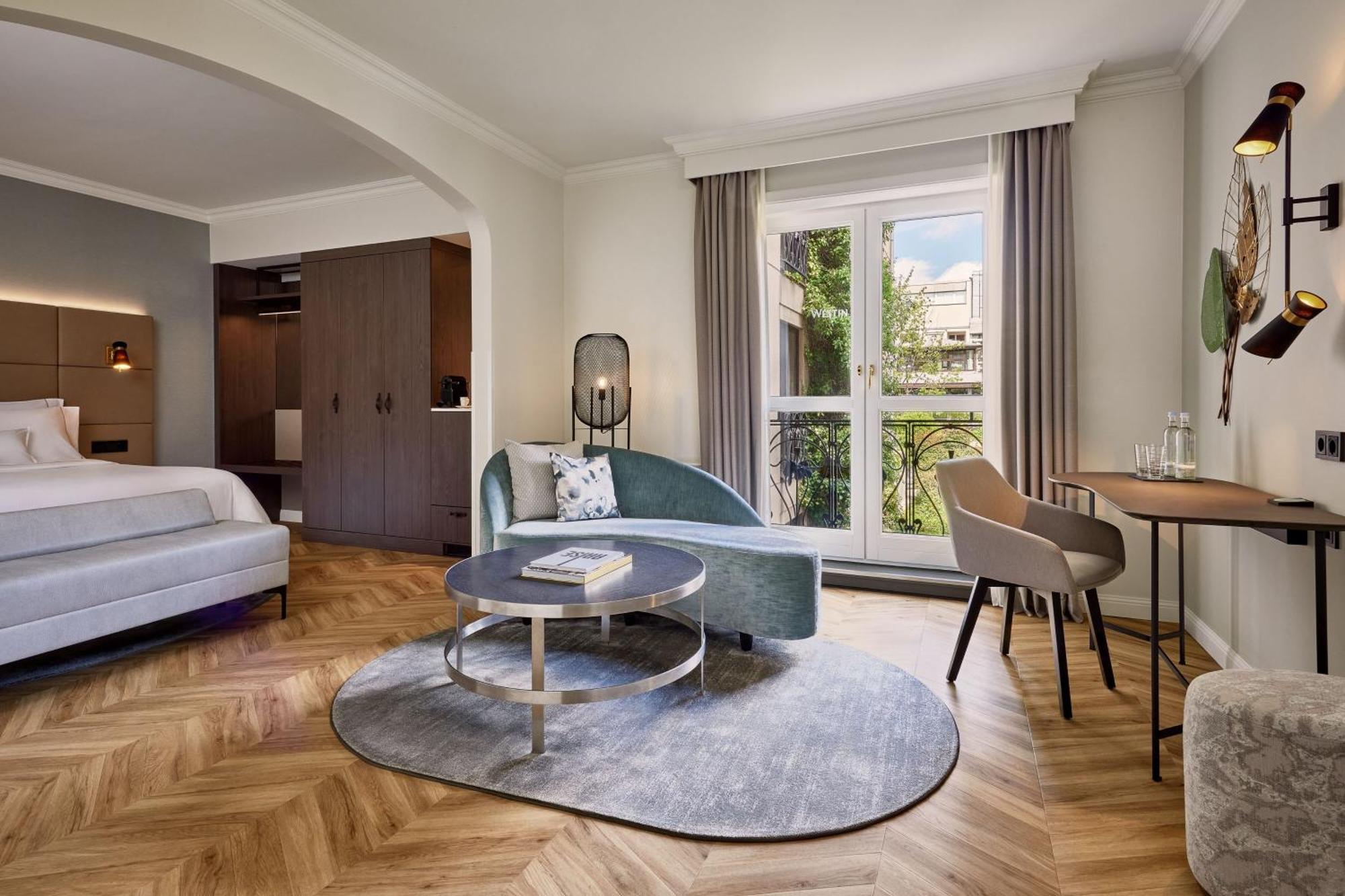 מלון The Westin Grand ברלין מראה חיצוני תמונה The photo shows a modern hotel room designed with a cozy and elegant aesthetic. In the foreground, there is a stylish, curved light blue sofa paired with a round coffee table. The table has a magazine or book on top. Nearby, there is a dining table w
