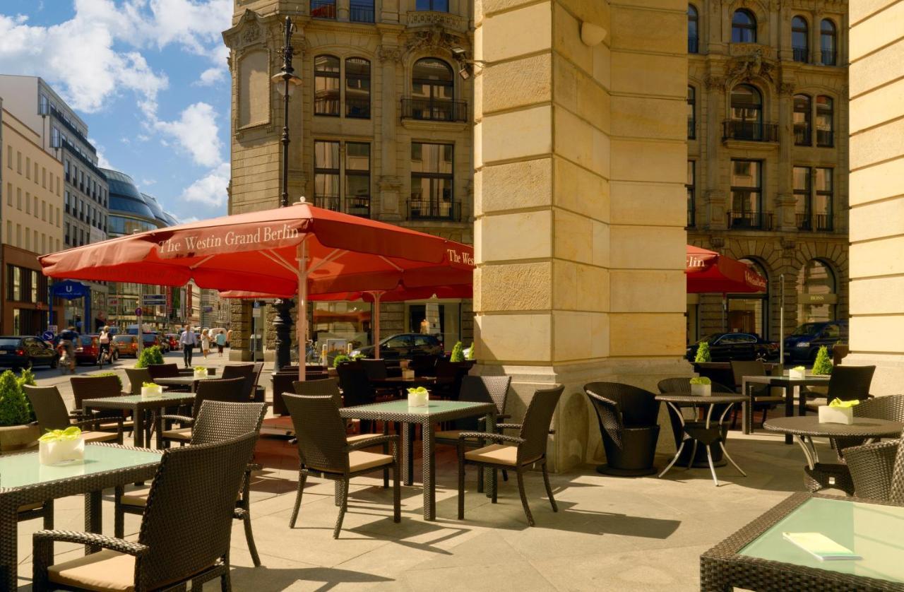 מלון The Westin Grand ברלין מראה חיצוני תמונה The photo shows an outdoor café area with several tables and chairs arranged under large red umbrellas. There are several potted plants on the tables, adding a touch of greenery. The café is situated in an urban setting, with impressive buildings in 